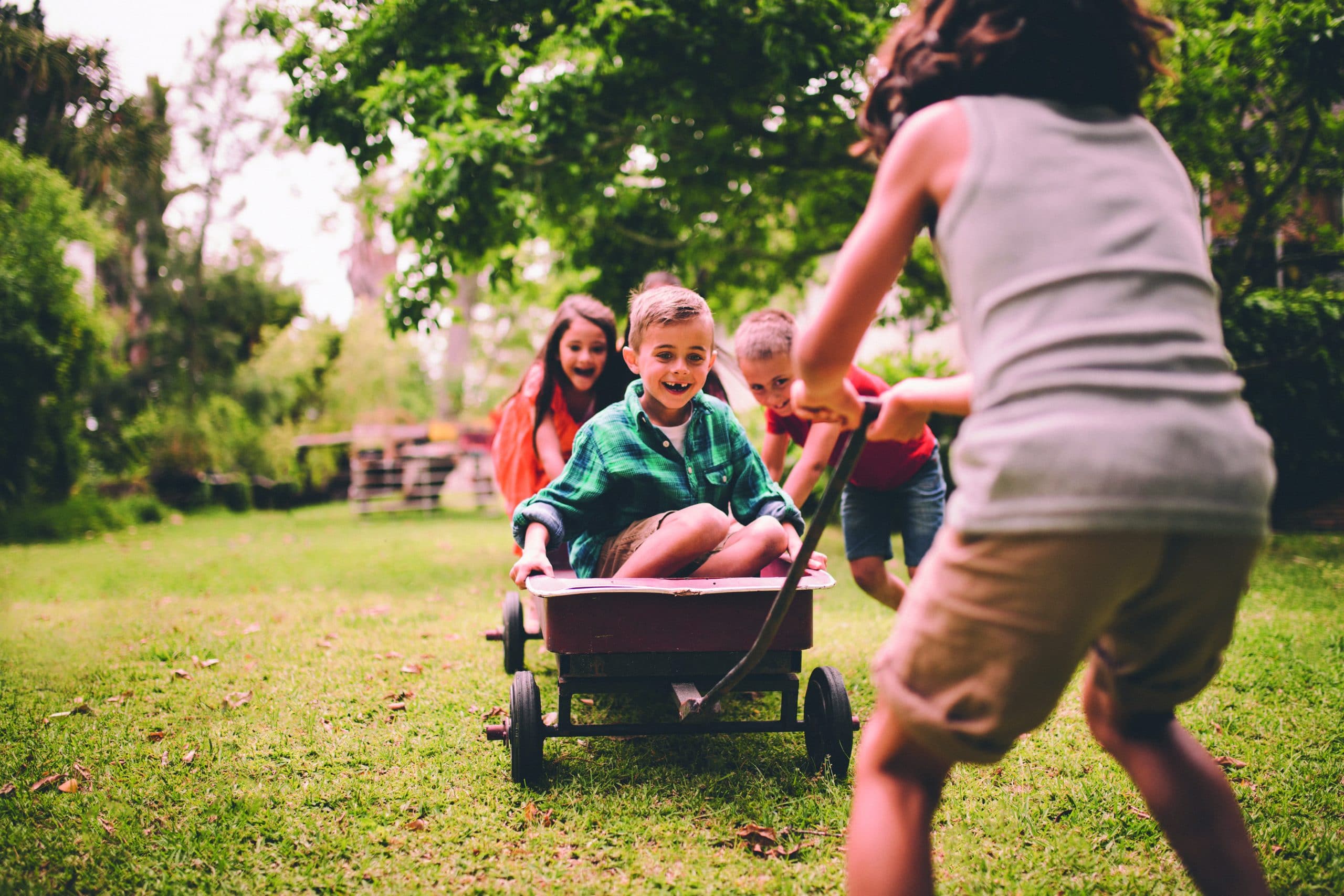 Ein Kind sitzt im Bollerwagen und wird von einem Kind gezogen und von anderen Kindern angeschoben - Quereinstieg als Erziehungskraft.